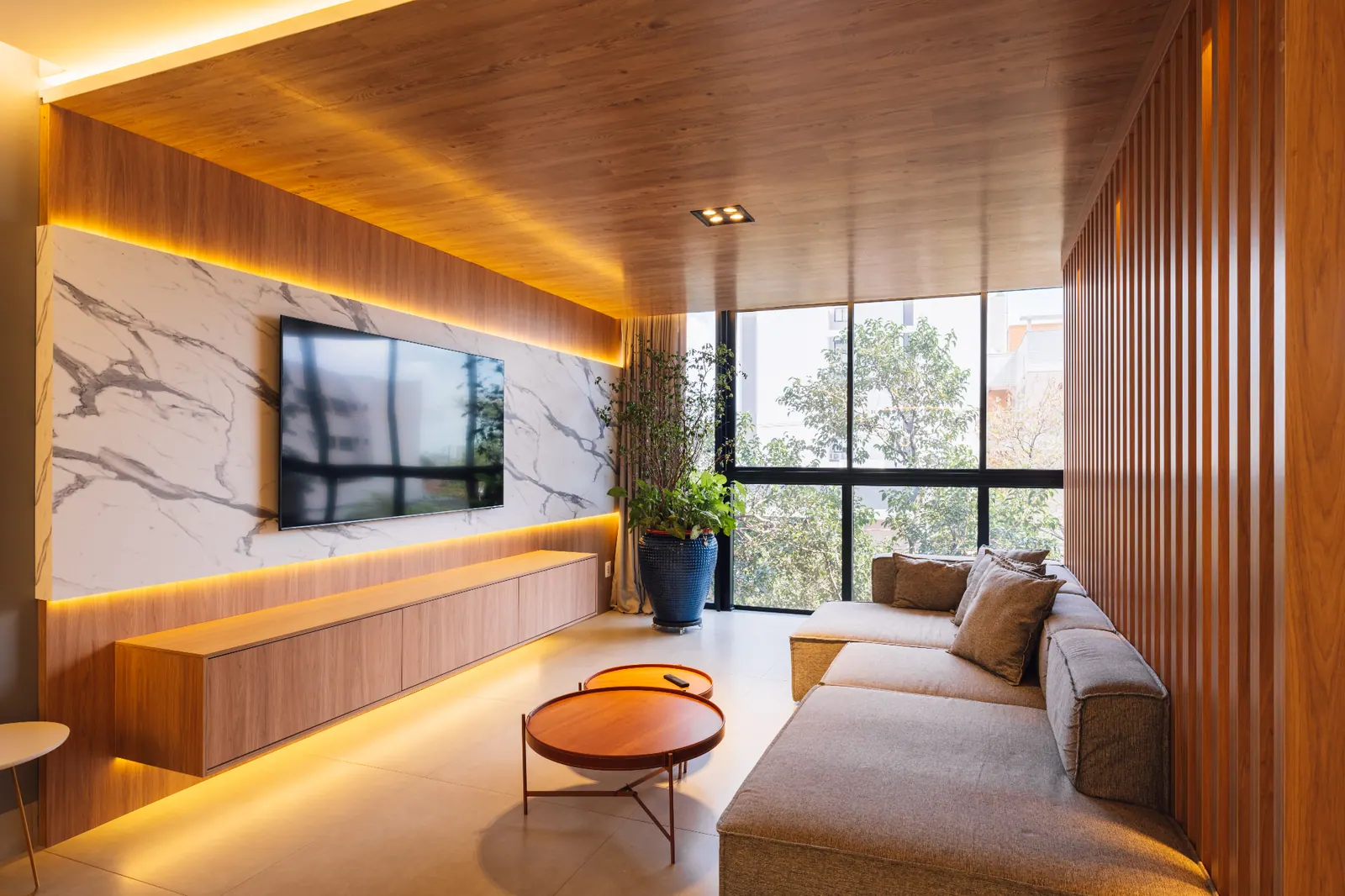 Living com painel de TV em mármore, forro em madeira e iluminação indireta